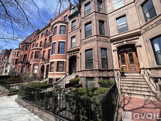 A row of brown brick townhouses with black metal fences and steps leading to the front doors.