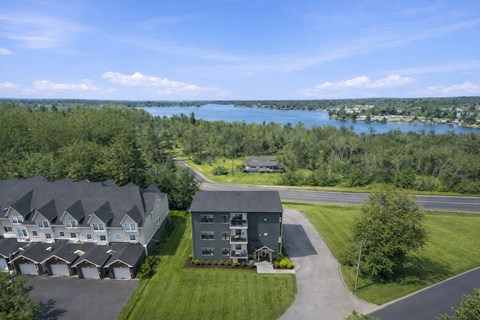 A large building with a grey roof is surrounded by a grassy area and a lake in the distance.