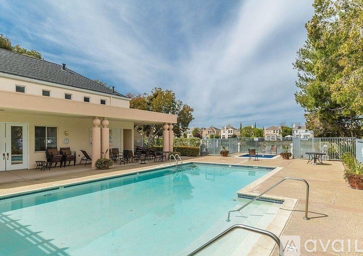 A swimming pool in front of a house with a patio and chairs.