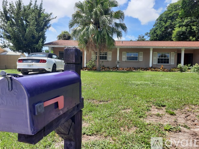 A purple mailbox is in the foreground with a white car and a house in the background.