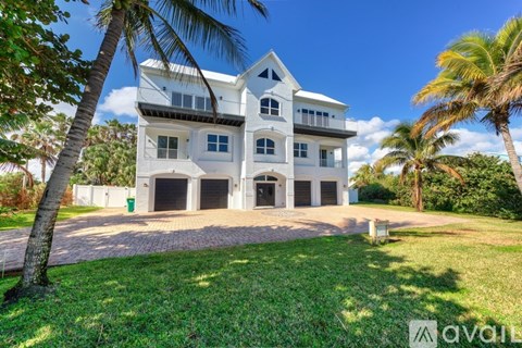 A large white house with a balcony on the second floor is surrounded by greenery and palm trees.