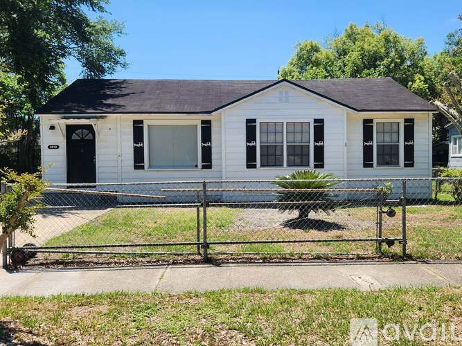 A small white house with a black door and windows is surrounded by a fence.