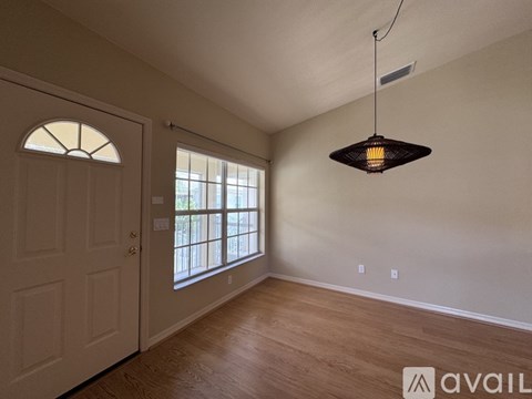 A room with a wooden floor, a white door, a window with a view of a fence and trees, and a hanging light fixture.