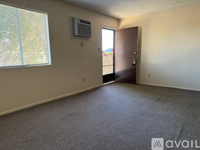 A room with a carpeted floor, a window with blinds, a wall-mounted air conditioner, and a door leading to a balcony.
