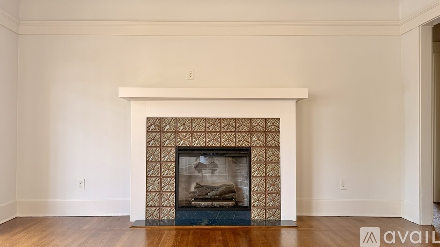 A fireplace with a patterned tile surround sits in a room with white walls and wood flooring.