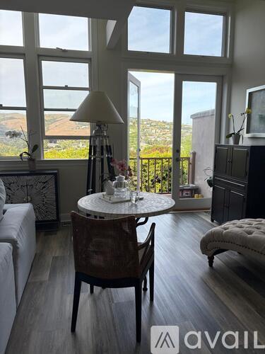 A living room with a white couch, a round table with a lamp, and a window overlooking a balcony.
