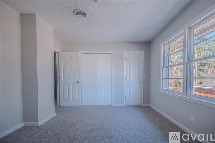 A room with a carpeted floor, a ceiling fan, and a window showing trees outside.