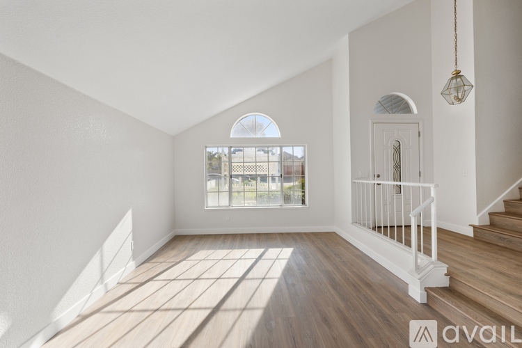A room with wooden floors and white walls, a staircase with white railings, and a large window with a view of a fence and trees.