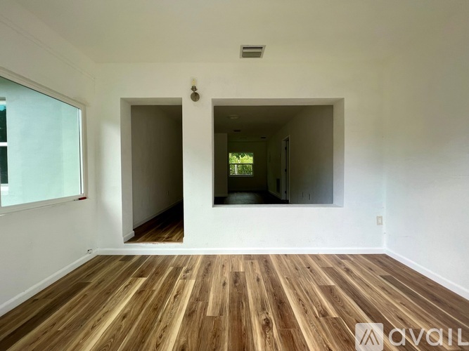 A room with wooden floors and white walls, with a window on the left and a doorway leading to another room.