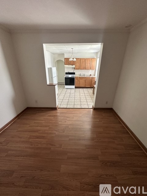 A room with wooden flooring and a kitchen area visible through the open door.