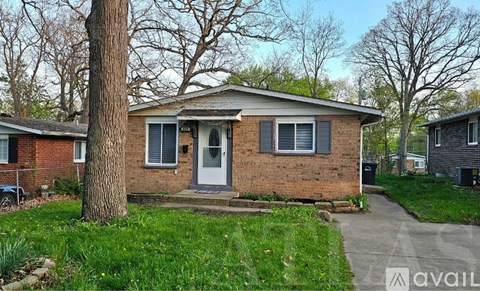 A small brick house with a white door and windows is surrounded by a grassy area and trees.