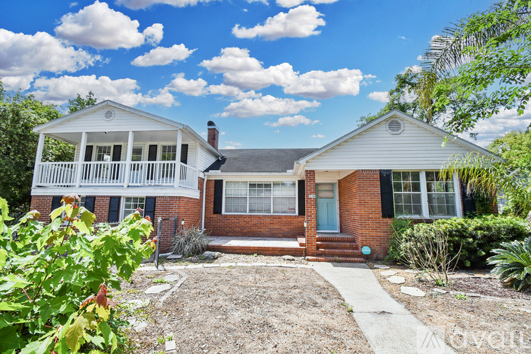 A house with a white porch and a brick exterior is surrounded by greenery.