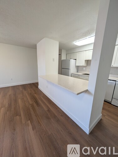 A room with wooden floors and white walls, with a kitchen area visible in the background.