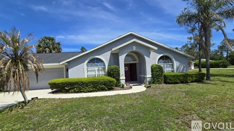A house with a grey roof and white walls is surrounded by greenery and has a clear blue sky above.