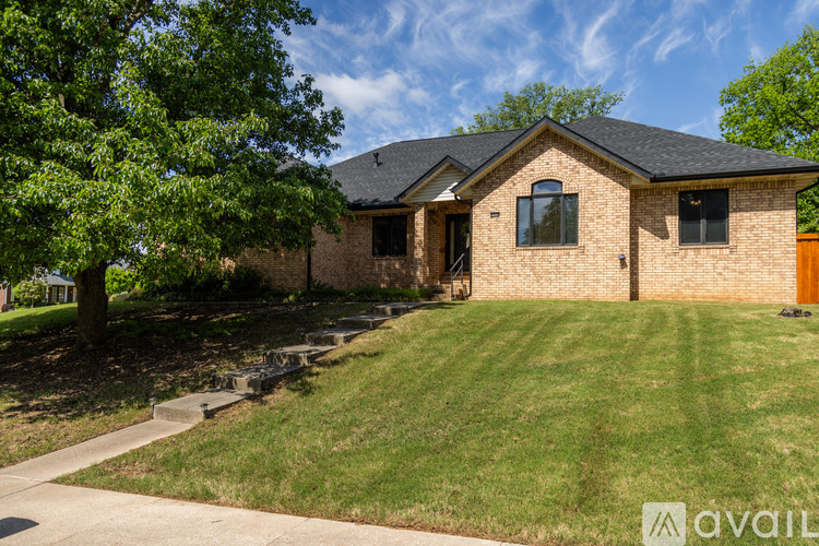 A house with a brown brick exterior and a black roof is surrounded by a green lawn and trees.