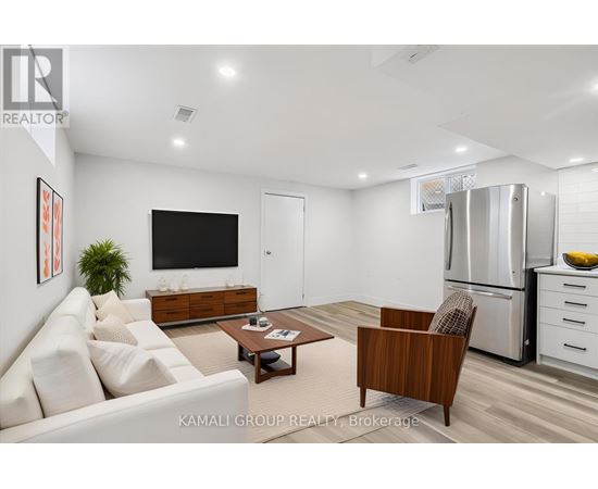A modern living room with a white sofa, a wooden armchair, a coffee table, a flat-screen TV, and a kitchen area with a refrigerator and a bowl of bananas.