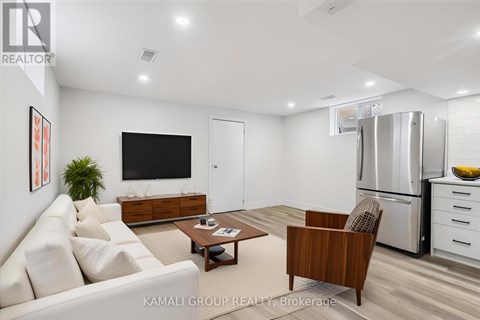 A modern living room with a white sofa, a wooden armchair, a coffee table, a flat-screen TV, and a kitchen area with a refrigerator and a bowl of bananas.