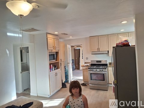 A girl in a white dress stands in a kitchen with a refrigerator to her right.