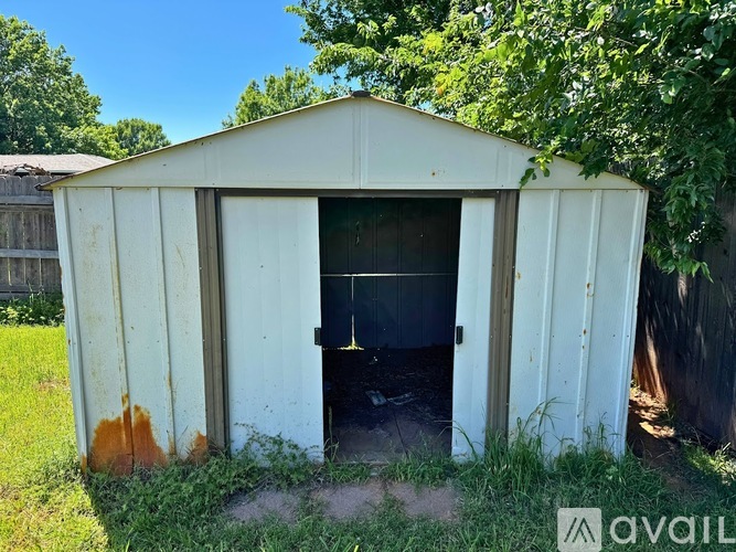 A shed with a white exterior is surrounded by greenery and has a door open to a dark interior.