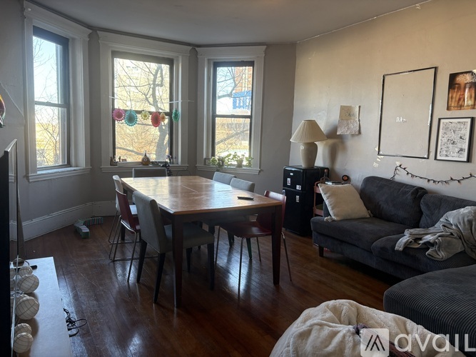 A living room with a grey couch, a wooden table and chairs, and a window with a view of trees.