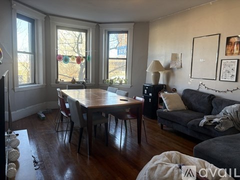 A living room with a grey couch, a wooden table and chairs, and a window with a view of trees.