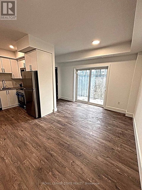A room with wood flooring and a kitchen area with a refrigerator.