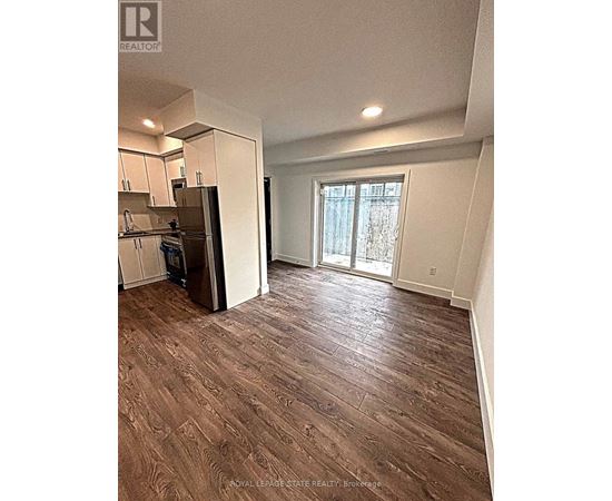 A room with wood flooring and a kitchen area with a refrigerator.