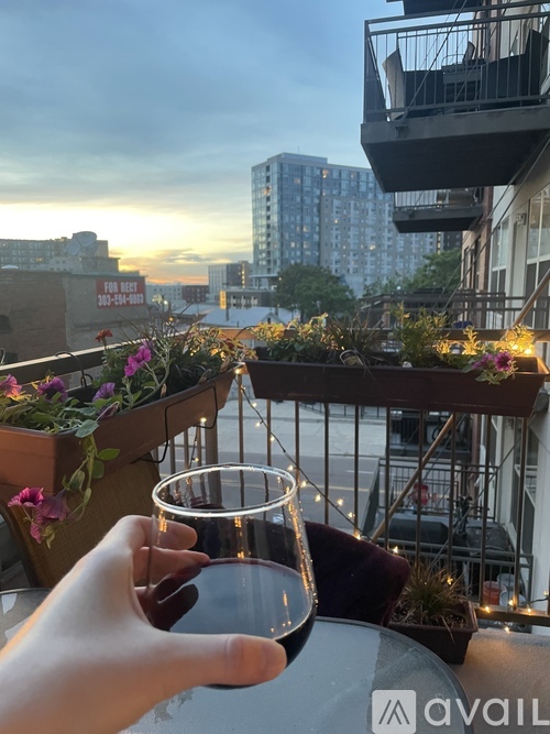 A person is holding a glass of wine on a balcony with flowers and buildings in the background.