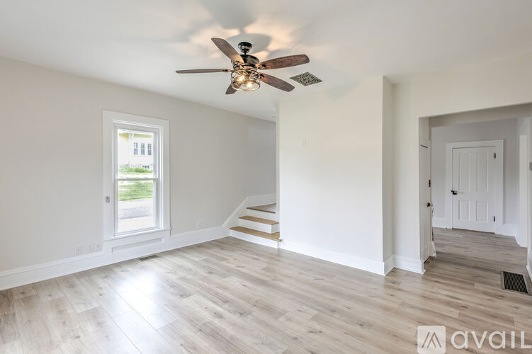 A room with a ceiling fan and light, hardwood floors, and a window with a view of a building outside.