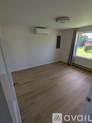 A room with wooden flooring and a window with a view of a red building and greenery outside.