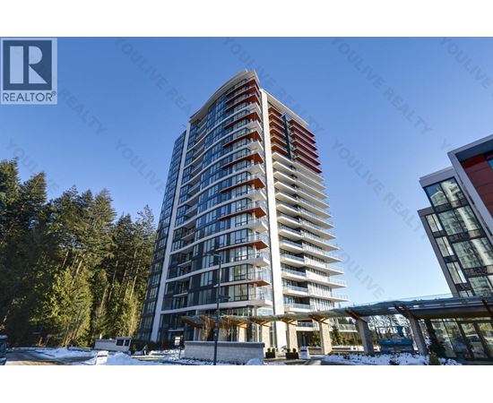 A tall residential building with balconies is surrounded by trees and has a clear blue sky in the background.