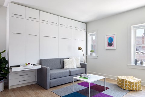 A grey couch with a white pillow and a grey blanket sits in a living room with a white cabinet and a window.