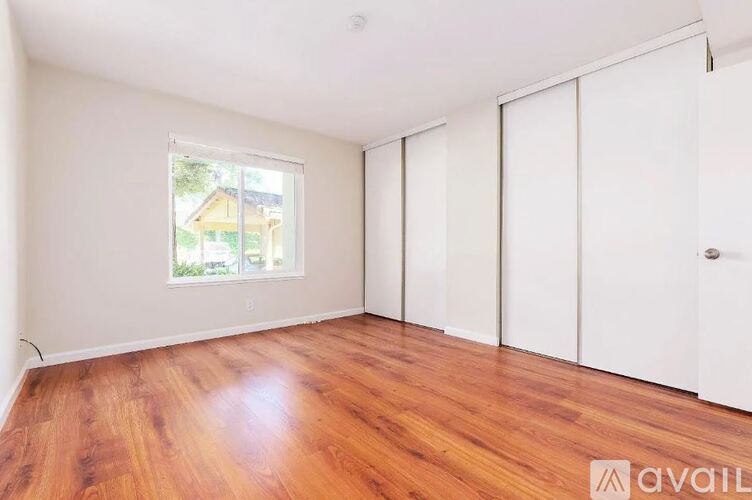 A room with wooden flooring and white walls, with a window showing a view of a house.