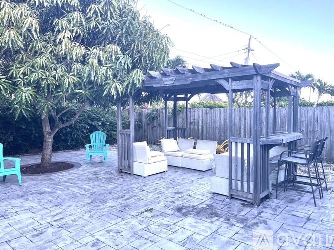 A wooden gazebo with white couches and chairs is surrounded by trees.