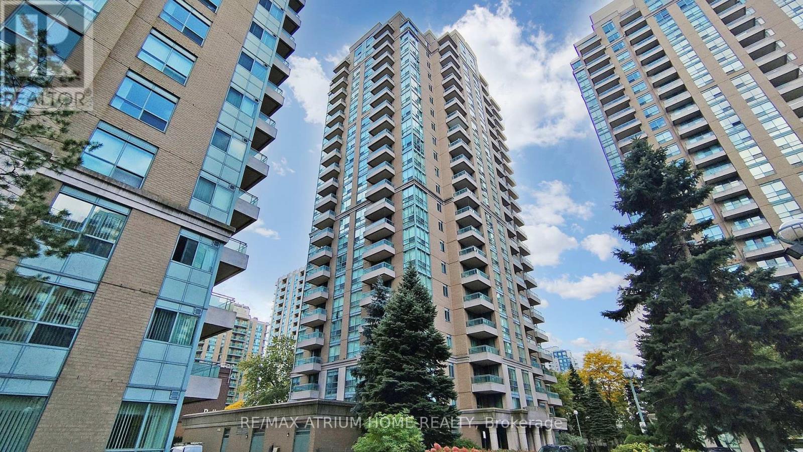 A tall residential building with balconies is surrounded by other buildings and trees.