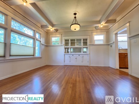 A spacious room with wooden floors and white walls, featuring a kitchenette with white cabinets and a window with blinds.