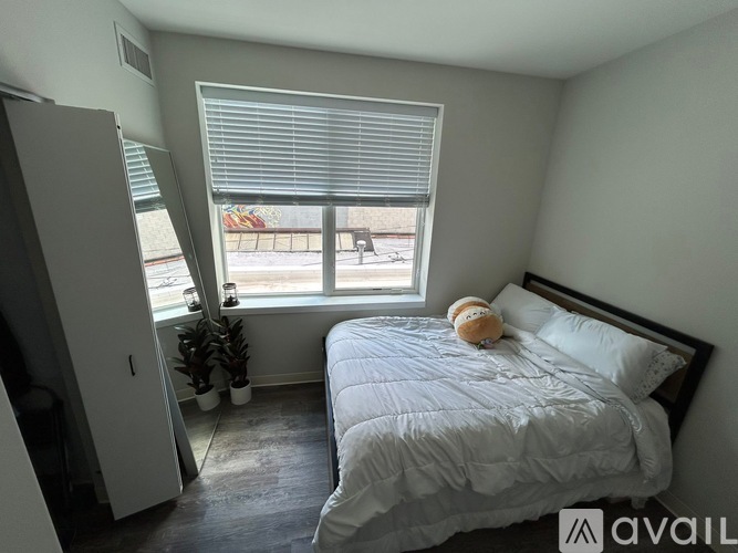 A bedroom with a bed, a window with blinds, a teddy bear on the bed, and a plant on the windowsill.