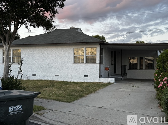 A house with a white exterior and a black roof is shown with a tree in front and a trash can to the left.