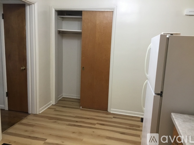 A room with a wooden floor and a white wall with a brown door and a white fridge.