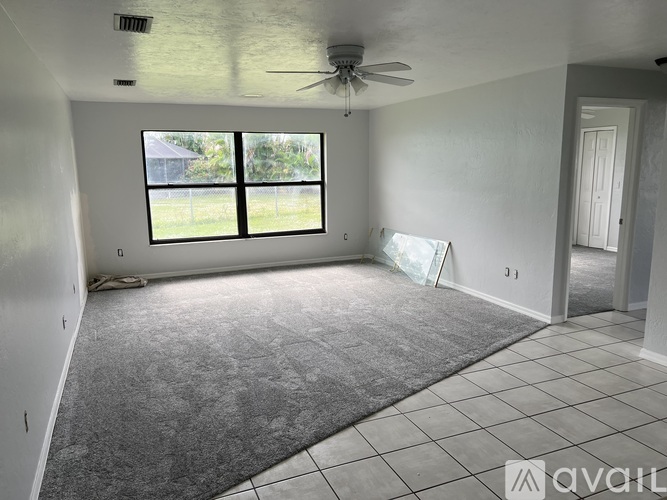 A room with a carpeted floor, a ceiling fan, and a window with a view of a house and trees outside.