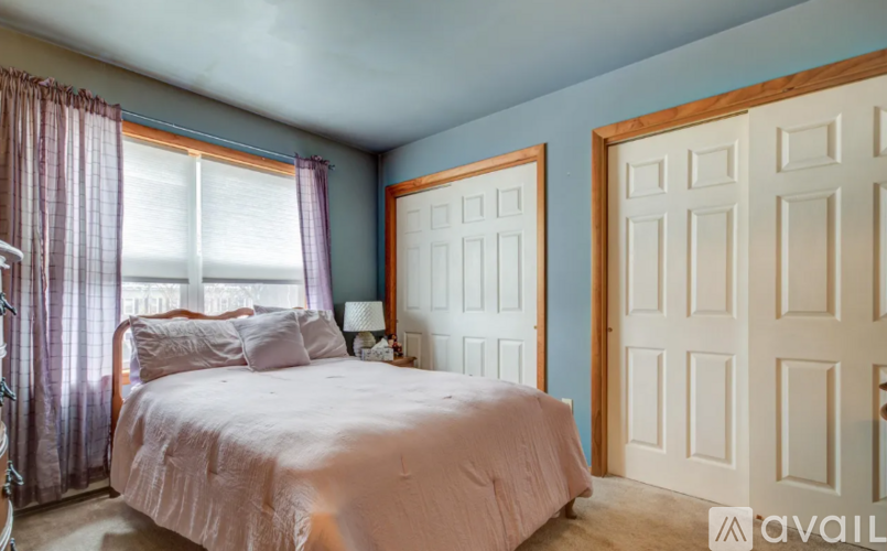 A bedroom with a bed, two pillows, a nightstand with a lamp, a window with curtains, and a door.