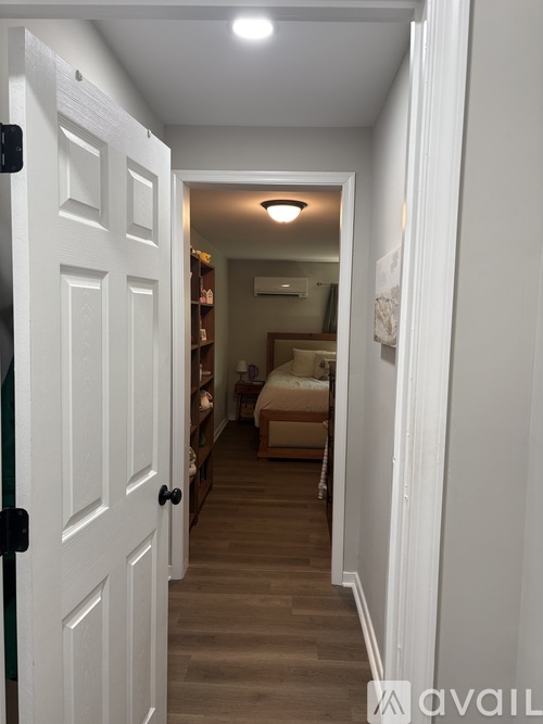 A hallway with a white door on the left and a bedroom with a bed and a couch in the distance.