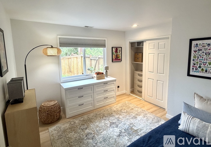 A bedroom with a bed, a window with blinds, a dresser with drawers, a wicker stool, a speaker, a rug, and a framed picture on the wall.