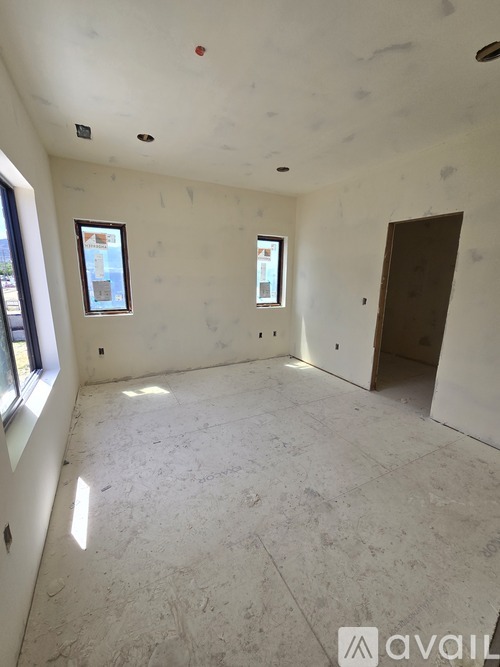 A room with a white ceiling and walls, with a window on the left wall and a door on the right wall.