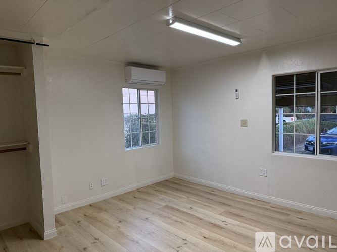 A room with wooden flooring and white walls, featuring a window with blinds and a wall-mounted air conditioner.