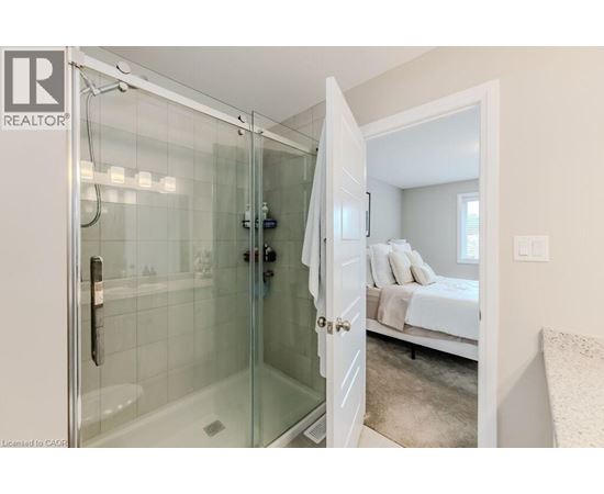 A glass shower stall with a white door is open to a bedroom with a bed and a window.