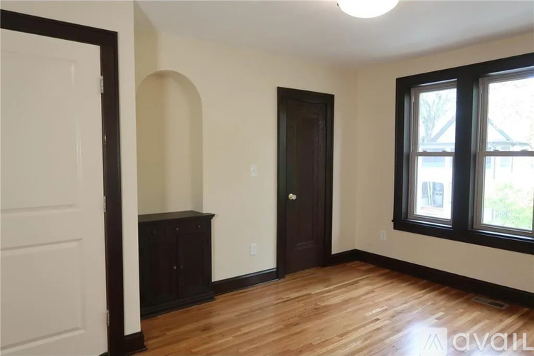 A room with a wooden floor, a white door, a brown door, and a window with a view of trees outside.