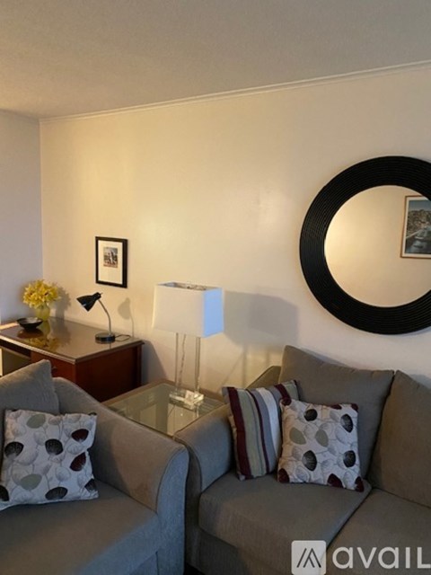 A living room with a grey couch, a black circular mirror, and a wooden table with a lamp and a vase of flowers.