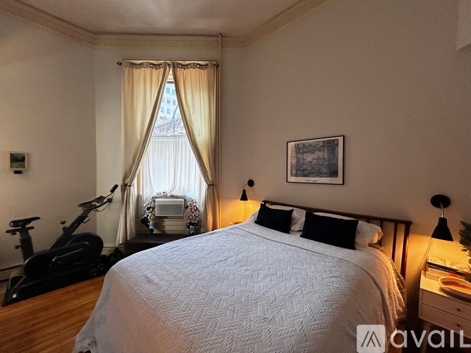 A bedroom with a large bed, a window with curtains, a picture on the wall, and a bicycle in the corner.