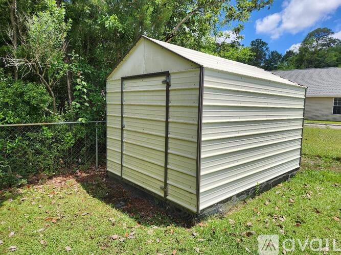 A small, white, metal shed with a black door sits in a grassy area.
